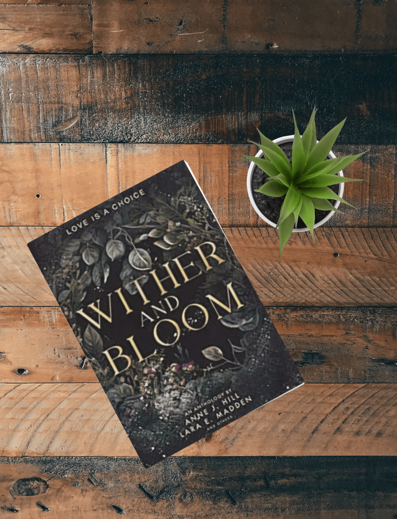 Wither and Bloom sits next to green plant on wooden table.