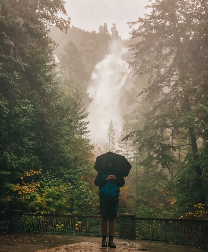 Nathaniel Luscombe stands with an umbrella in front of a waterfall. 