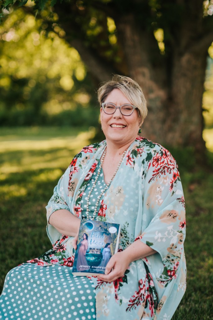 Author Jenny Knipfer smiles while holding one of her books outside. 